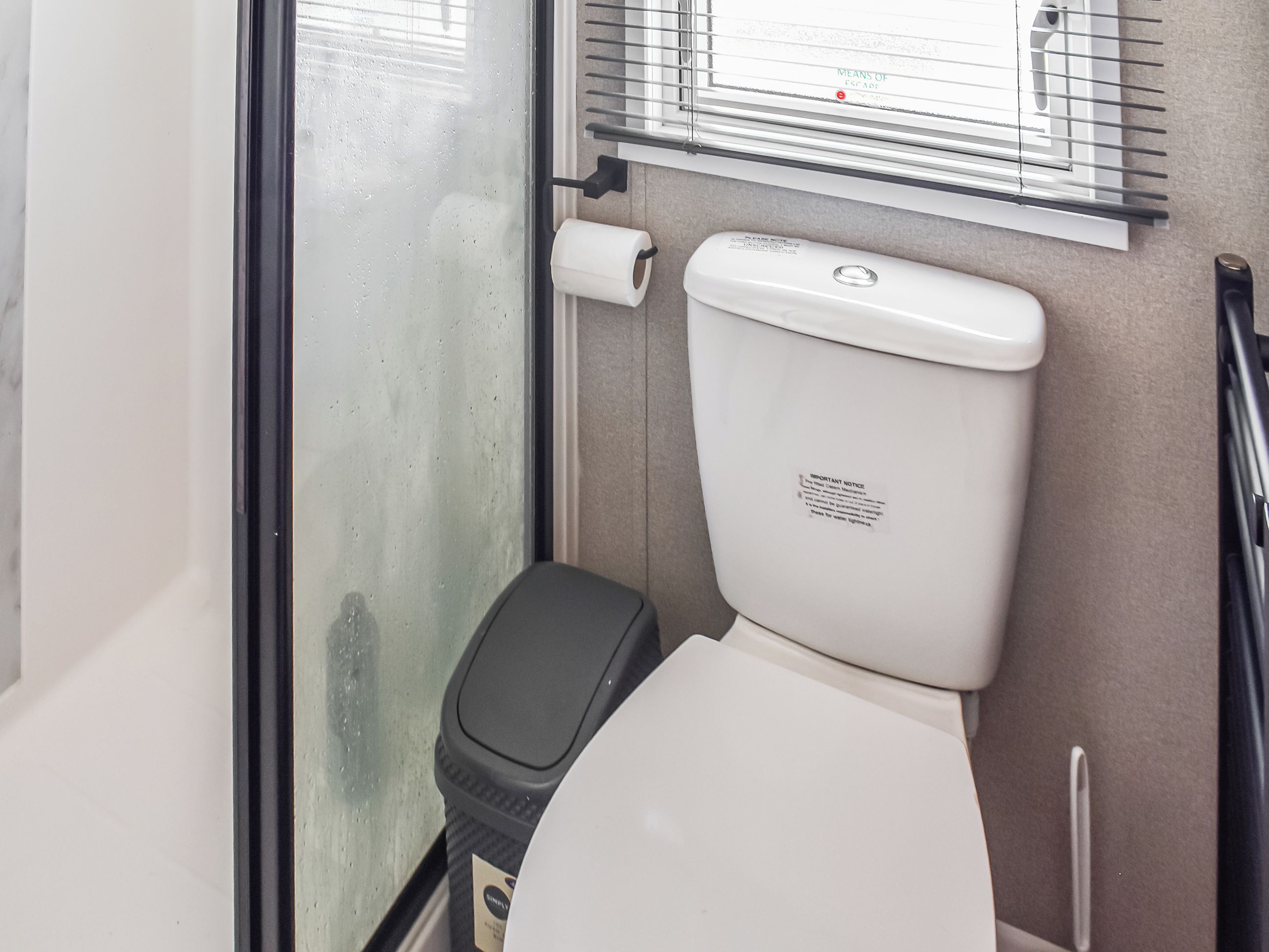 A compact bathroom with a toilet, a small trash bin, and a window with blinds.