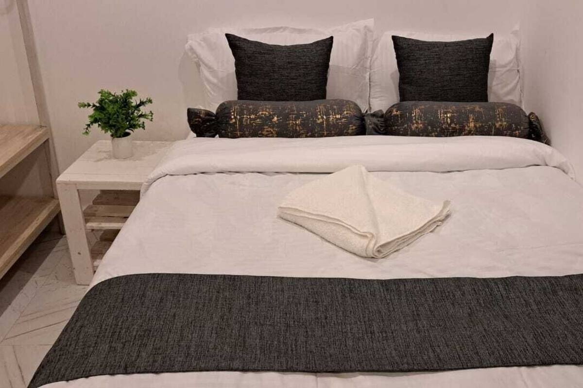Un lit bien fait, avec une literie blanche et gris foncé, une petite table de chevet ornée d’une plante et une couverture grise.