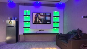 Una sala de estar moderna con un televisor de pantalla plana montado en la pared, iluminación verde, un refrigerador, un sofá y un zapatero.