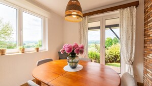 Comedor con una mesa redonda de madera, sillas, un jarrón con flores, una lámpara colgante y vistas a través de una puerta de cristal.
