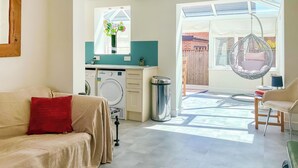Una sala de estar con un sofá color beige, un cojín rojo, una lavadora y una silla colgante.