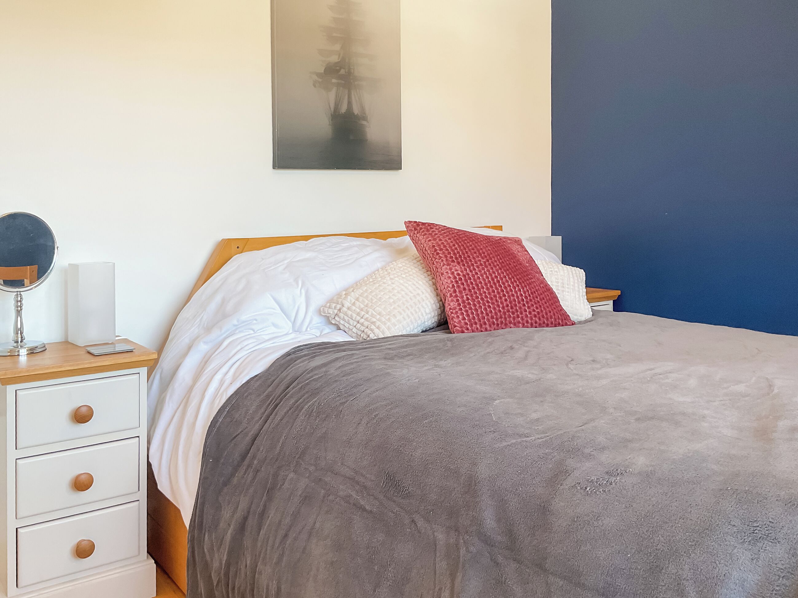 Un lit bien fait, avec une couverture grise, des oreillers blancs et un coussin rouge. On y trouve une table de chevet avec une lampe, un miroir et une petite boîte blanche. Une table de nuit en bois se trouve à gauche du lit.