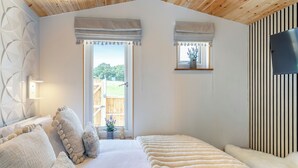 A bedroom with a large bed, two windows with curtains, a wooden ceiling, and a mounted TV. - Barn Owl Lodge - UK49903 (Dalton, near Wigan)