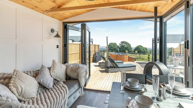 A cozy room with a wooden ceiling, a sofa with fluffy pillows, a dining table set with plates and glasses, and a hot tub in the background. - Barn Owl Lodge - UK49903 (Dalton, near Wigan)