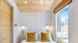 Ett sovrum med ett träpanelat tak, en väggpanel med geometriskt mönster, en säng med ett gult täcke och ett fönster med utsikt över naturen.