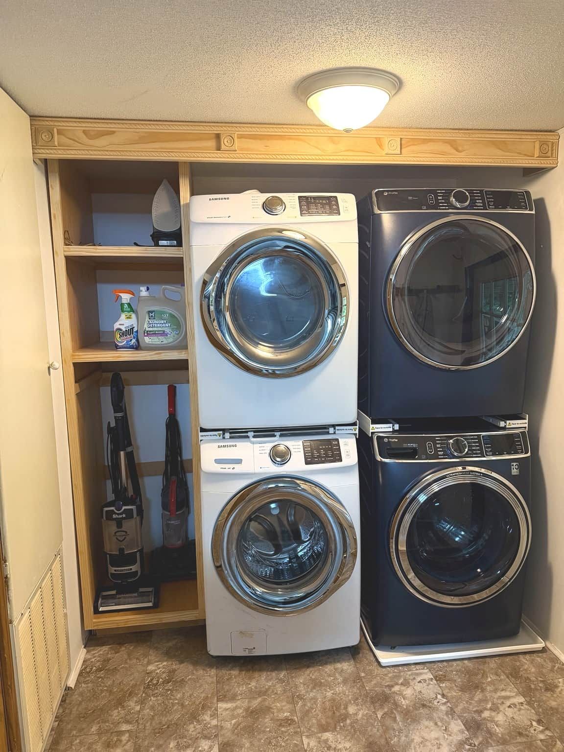 Un cuarto de lavandería con dos lavadoras de carga frontal, un estante con productos de limpieza y una aspiradora.