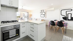 Een moderne keuken met witte kastjes, een gasfornuis en een ingebouwde oven. Er is een eethoek met een tafel en stoelen, en abstracte kunst aan de muren.