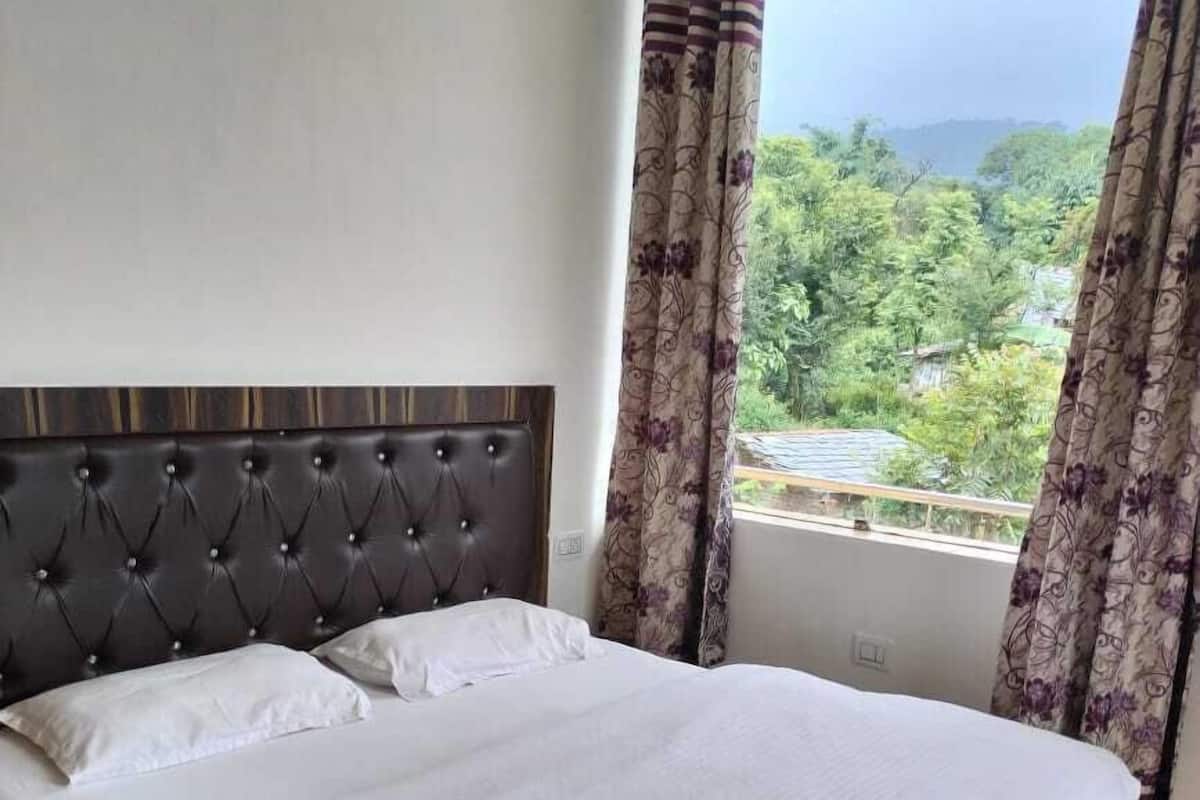 A double bed with a tufted headboard, white bedding, and a view of greenery through a window with floral curtains.