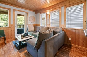 A cozy room with a wooden ceiling and walls, a grey sofa with cushions, a small table with a laptop, and a door leading outside.