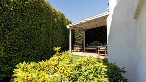 A covered patio area with outdoor furniture, surrounded by well-maintained hedges and a white wall.