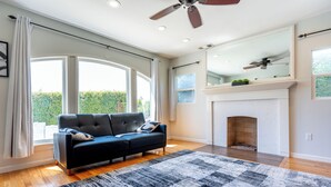 A modern living room with a black sofa, a patterned rug, a fireplace, and large windows offering a view of greenery.