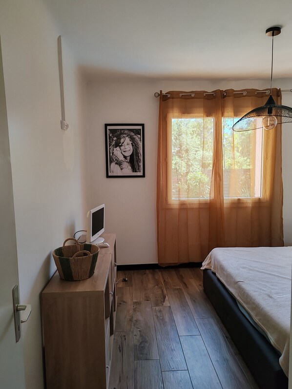 Ein Schlafzimmer mit einem Bett, einem Schreibtisch mit Computer, einem Fenster mit Vorhängen und einem gerahmten Bild an der Wand.