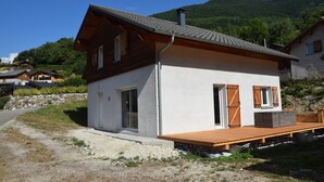 A two-story house with a sloped roof, a wooden deck, and a stone wall in front. - Family home in the heart of the Tarentaise (Moûtiers)