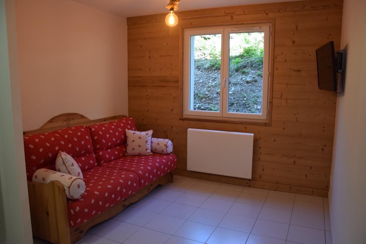 A cozy room with a wooden wall, a red sofa with floral cushions, a window with a view of greenery, and a white radiator.