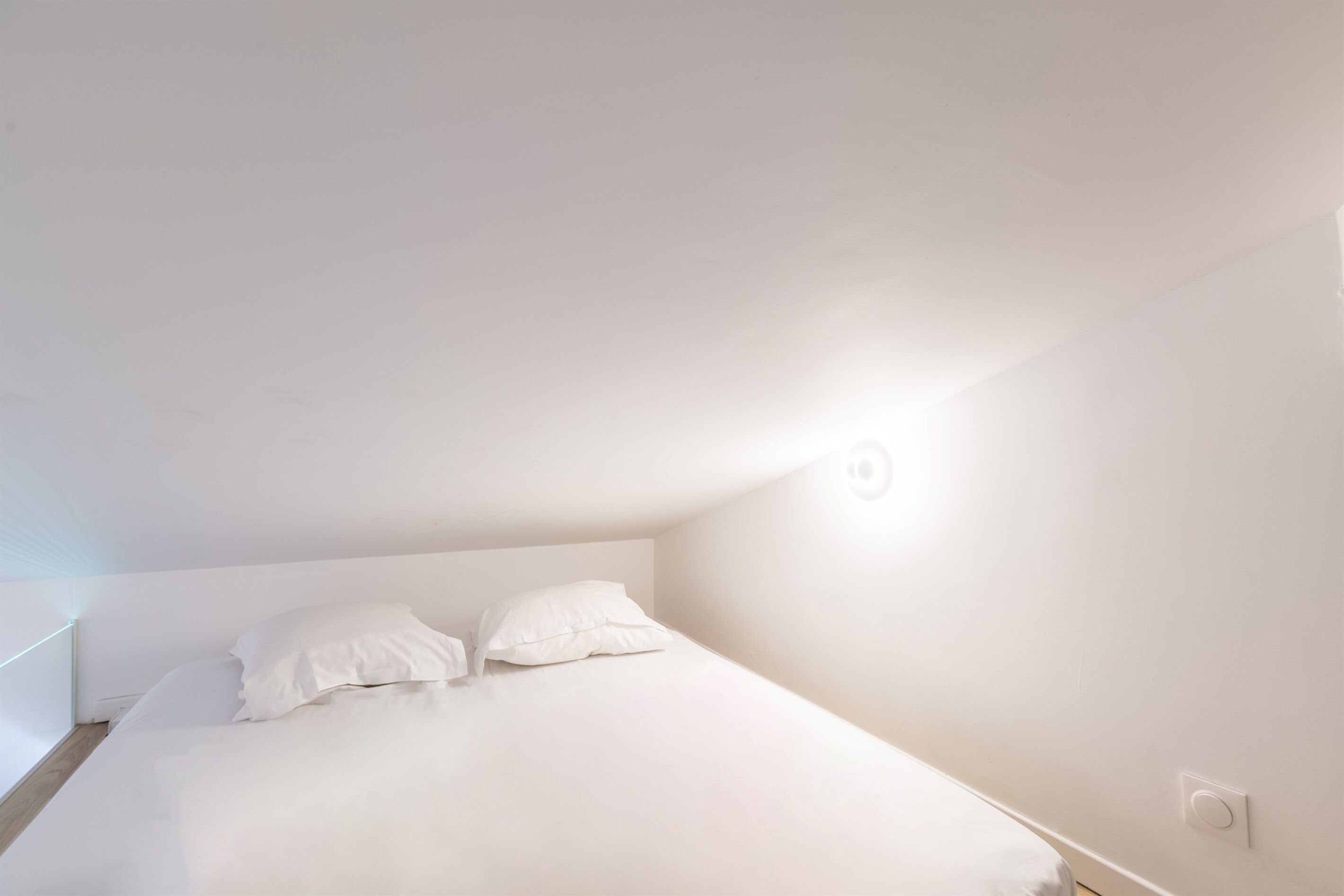 A small, white, single-bed room with a slanted ceiling and a single wall-mounted light fixture.