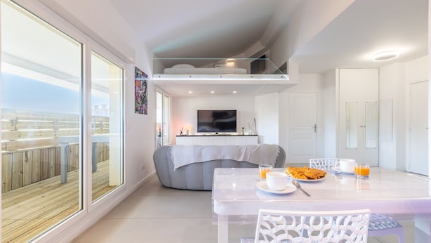 A modern living room with a dining area, a sofa, a TV, and a balcony with a wooden deck.