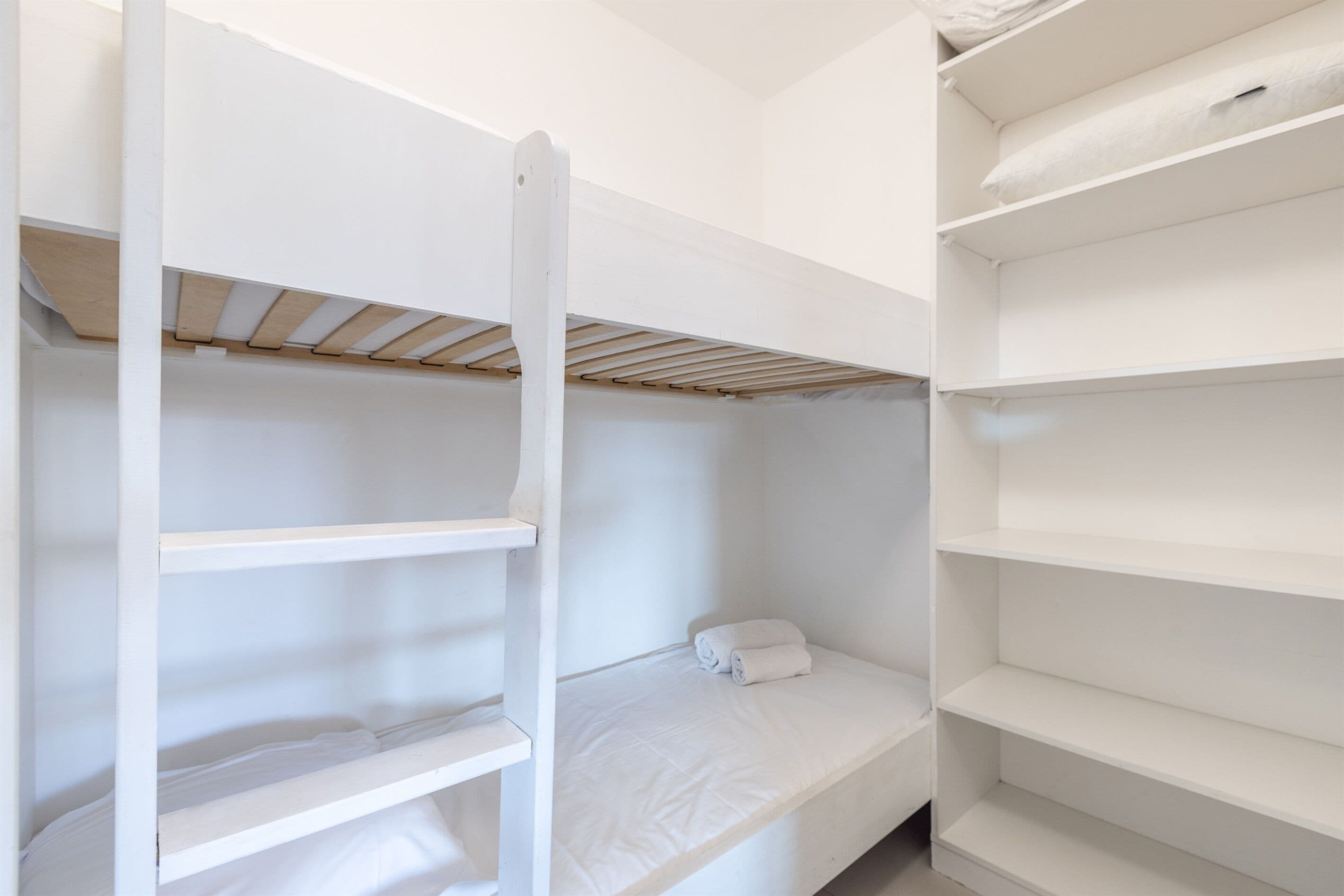 Un lit superposé avec une échelle en bois, un lit simple avec une couverture blanche enroulée, et une étagère blanche sur laquelle repose un oreiller.