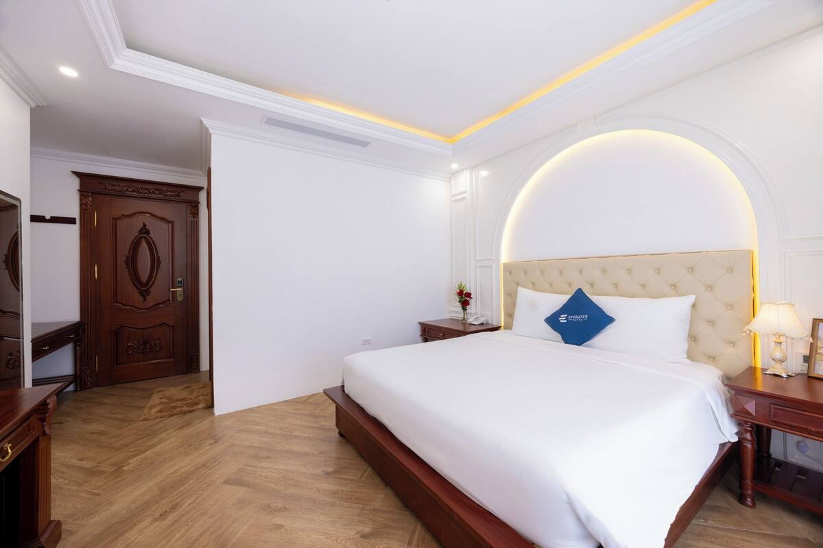 A hotel room with a large bed, wooden floor, a wooden wardrobe, a nightstand with a lamp, and a mirror with a curved frame.