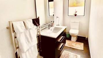 Un baño moderno con un espejo grande, un lavamanos y un inodoro. Hay un perchero con toallas, una alfombra en el suelo y un cuadro en la pared.