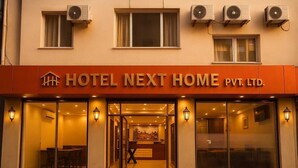 L’entrée d’un hôtel, avec une enseigne indiquant « HOTEL NEXT HOME PVT. LTD. » et plusieurs unités de climatisation fixées à la façade.