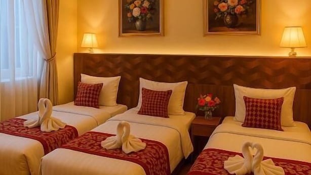 Une chambre d’hôtel avec trois lits, chacun recouvert d’une couverture à carreaux rouges et blancs, et surmonté d’une serviette blanche disposée en forme de cygne. De chaque côté des lits, on trouve deux lampes fixées au mur, et au-dessus des lits, deux tableaux encadrés représentant des motifs floraux.