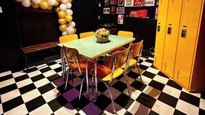 Una habitación con suelo de cuadros, una mesa con sillas, un banco, taquillas amarillas y una pared decorada con diversos carteles.