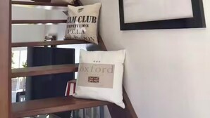 Un escalier avec une rampe en bois, un mur blanc orné de photographies en noir et blanc encadrées, et deux coussins avec des motifs textuels et des drapeaux.