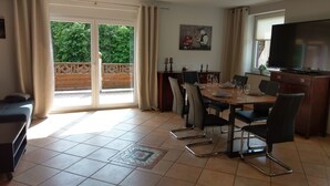 Een eethoek met een tafel voor vier personen, stoelen, een televisie en uitzicht op groen buiten.