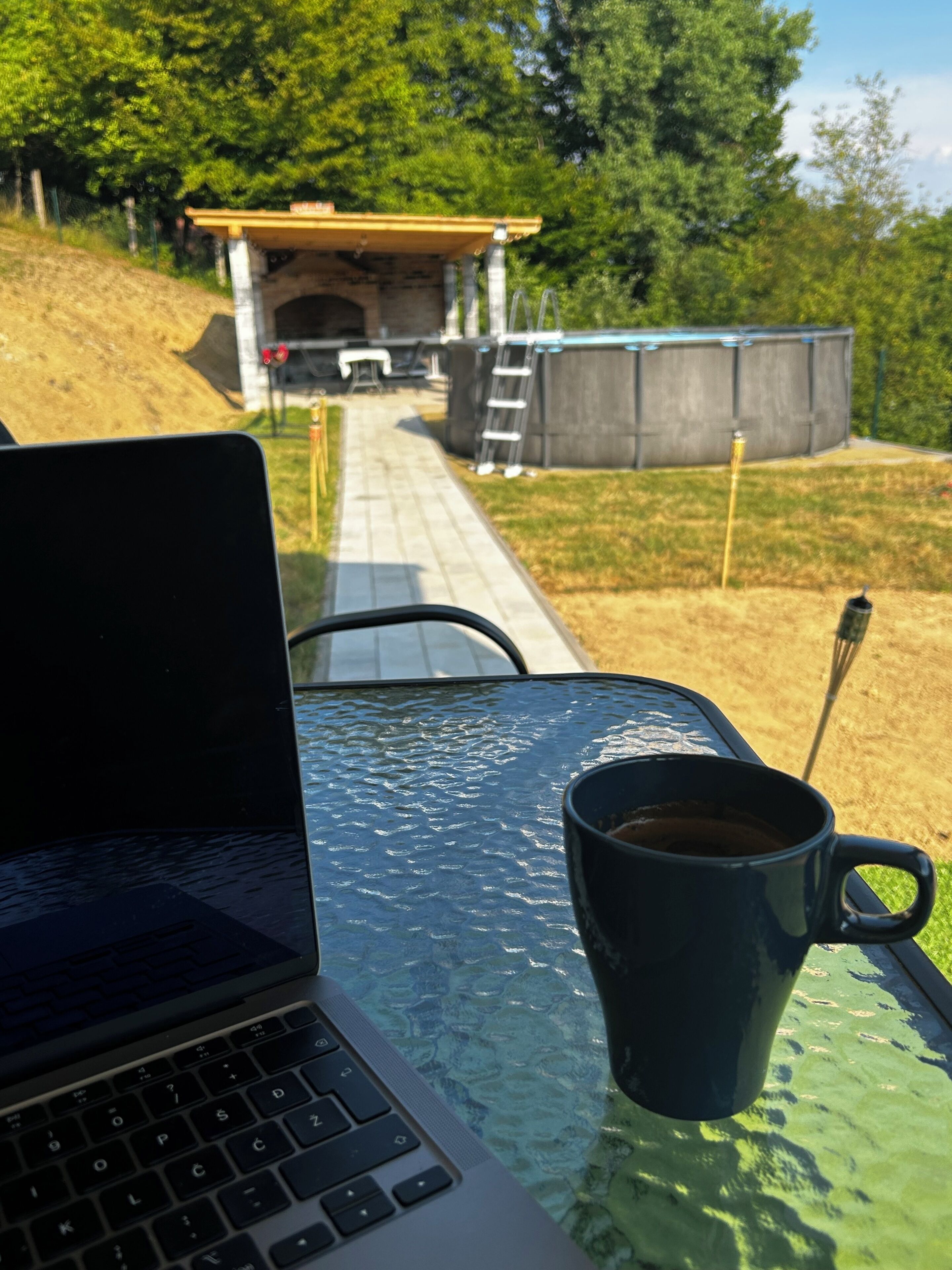 A laptop on a table with a black mug, a clear glass table, and a view of a pathway leading to a building with a covered area and a ladder.