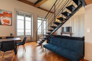 A modern living room with a wooden herringbone floor, a blue sofa, a dining area with a wooden table and chairs, and a black spiral staircase. - Smartstay - Vue panoramique sur Lyon et la saone (Lyon)