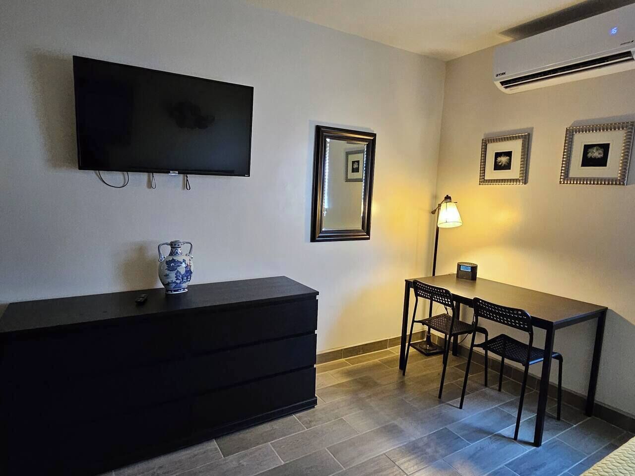 Une pièce dotée d’un téléviseur à écran plat fixé au mur, d’une petite table avec deux chaises, d’une lampe et d’un vase décoratif posé sur une commode.
