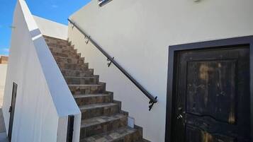 Un escalier moderne avec des marches en pierre menant à une porte en bois sombre, le tout adossé à un mur blanc sur lequel est fixée une applique lumineuse.