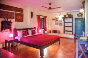 A bedroom with a bed, bedside tables, a ceiling fan, a window with curtains, a wall-mounted shelf, and a door. - Yuppi Hippi Hideout (Anjuna)