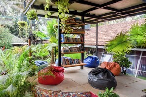A covered outdoor area with a bookshelf, bean bags, and various plants. - Yuppi Hippi Hideout (Anjuna)