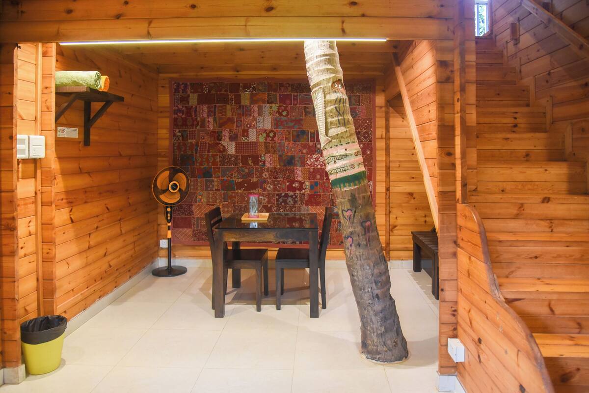 A wooden cabin interior with a dining table, chairs, a fan, a quilted wall, and a wooden staircase.