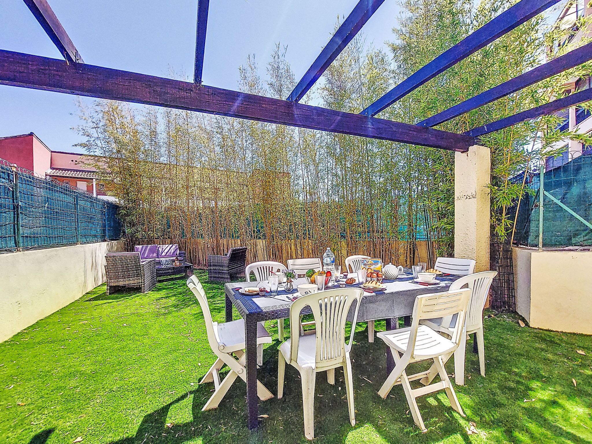 An outdoor dining area with a table set for six, surrounded by white plastic chairs, under a blue metal structure.