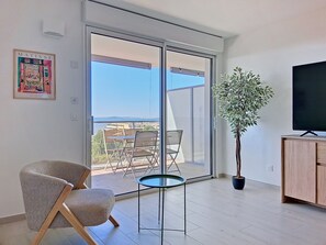 A modern living room with a sliding glass door leading to a balcony, a television, a potted plant, and a framed artwork.