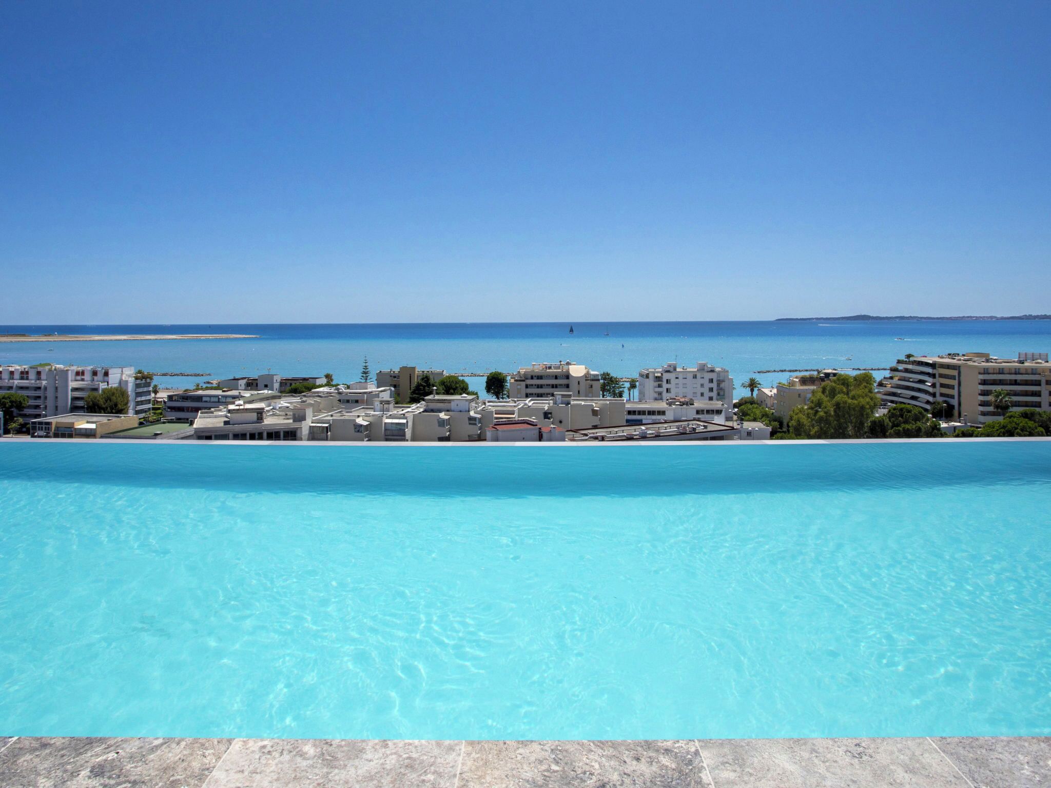 A pool with clear blue water, surrounded by buildings with a view of the sea.