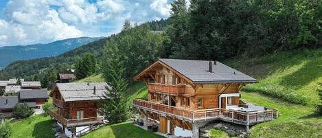 A wooden chalet with a balcony, surrounded by lush greenery and mountains in the background.