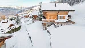 A snow-covered wooden chalet with a balcony, surrounded by snow-covered trees and mountains in the background.