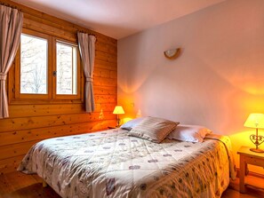 Un dormitorio acogedor con suelo de madera, una cama con ropa de cama floral, dos lámparas de noche y un ventanal con cortinas.