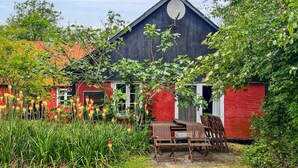 Een rode schuur met een zwart dak, omgeven door groen en een klein terras met houten stoelen.