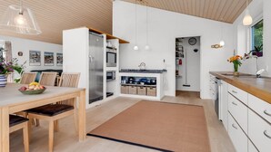 Eine moderne Küche mit weißen Schränken, einer Holzausziehtafel und einem eingebauten Backofen. Ein Essbereich mit Tisch und Stühlen sowie eine Kücheninsel mit Spüle und Arbeitsfläche sind ebenfalls vorhanden.