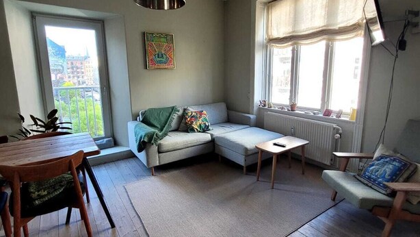 A spacious living room with a wooden floor, a sofa, a coffee table, and a dining table with chairs. There is a large window with a view of buildings outside. - Best location at Vesterbro and close to subway (Copenhagen v)