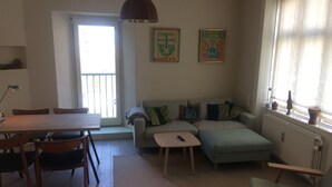 A living room with a sofa, a coffee table, and a dining table with chairs. There is a window with curtains and a radiator. - Best location at Vesterbro and close to subway (Copenhagen v)