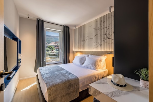 A hotel room with a bed, a TV, a window with curtains, a wall-mounted lamp, and a plant. - Beachfront Penthouse Residence (Podstrana)
