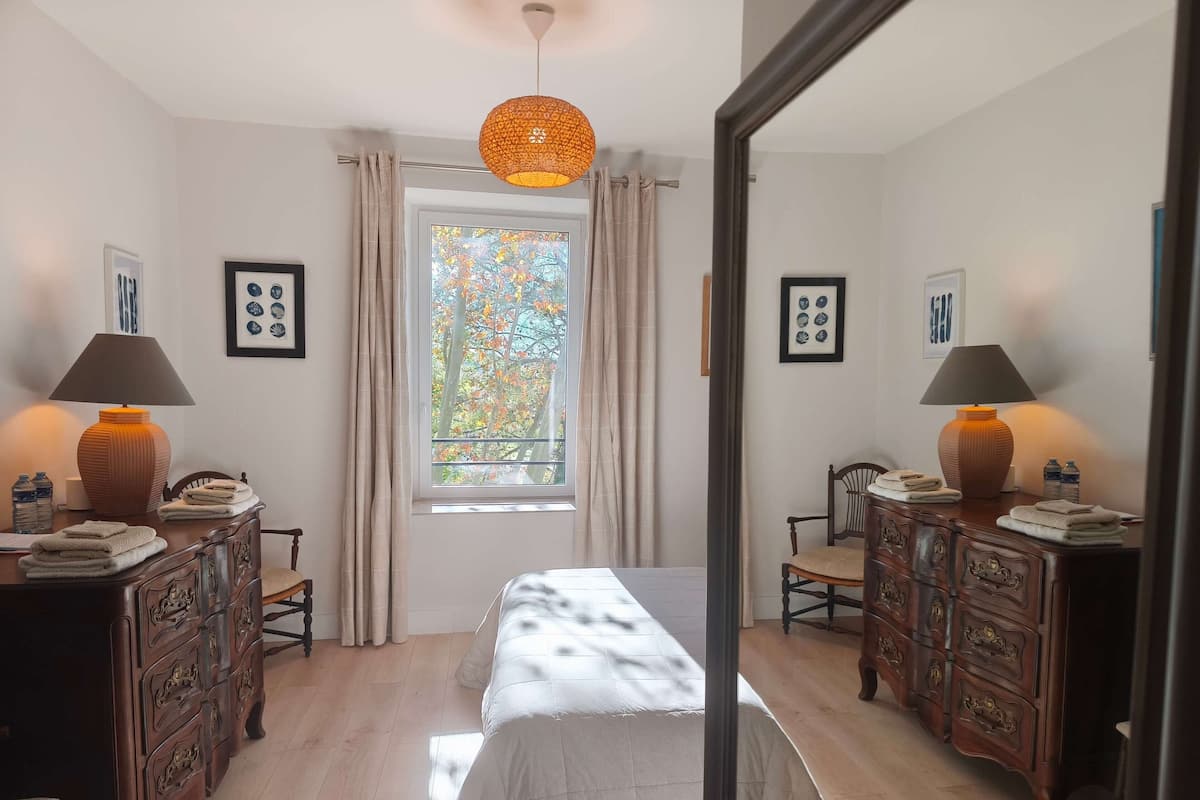 A bedroom with a bed, two nightstands with lamps, a dresser, a window with curtains, and a mirror.