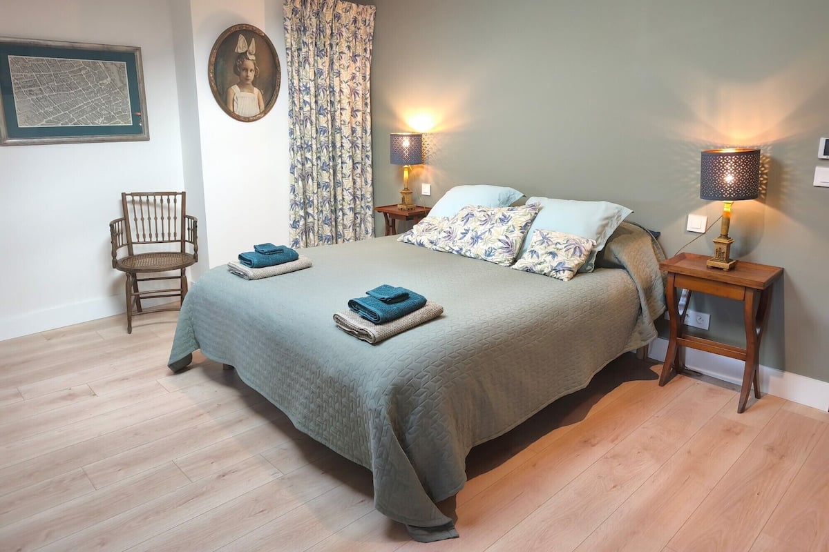 A neatly made bed with a green bedspread and two pillows, flanked by two bedside tables with lamps, a chair, and a framed map on the wall.