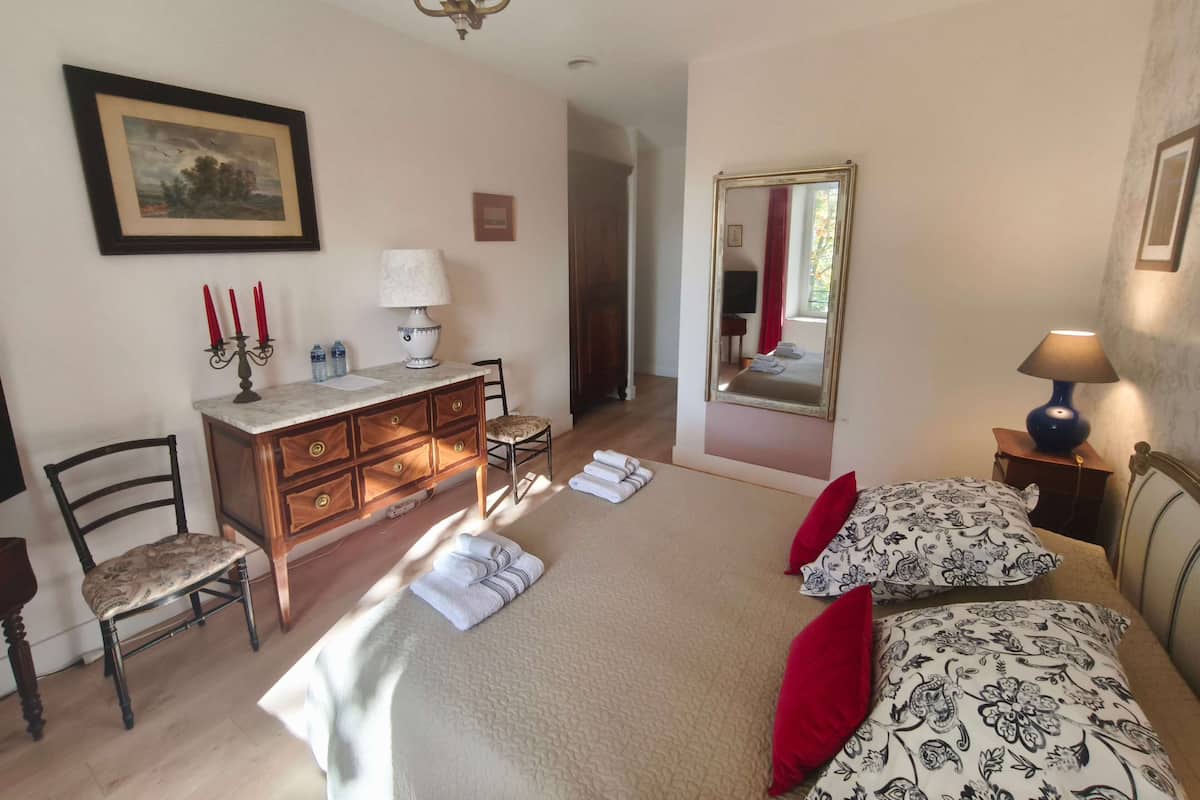 A bedroom with a bed, a dresser, a chair, a desk, a lamp, a mirror, and a chandelier.