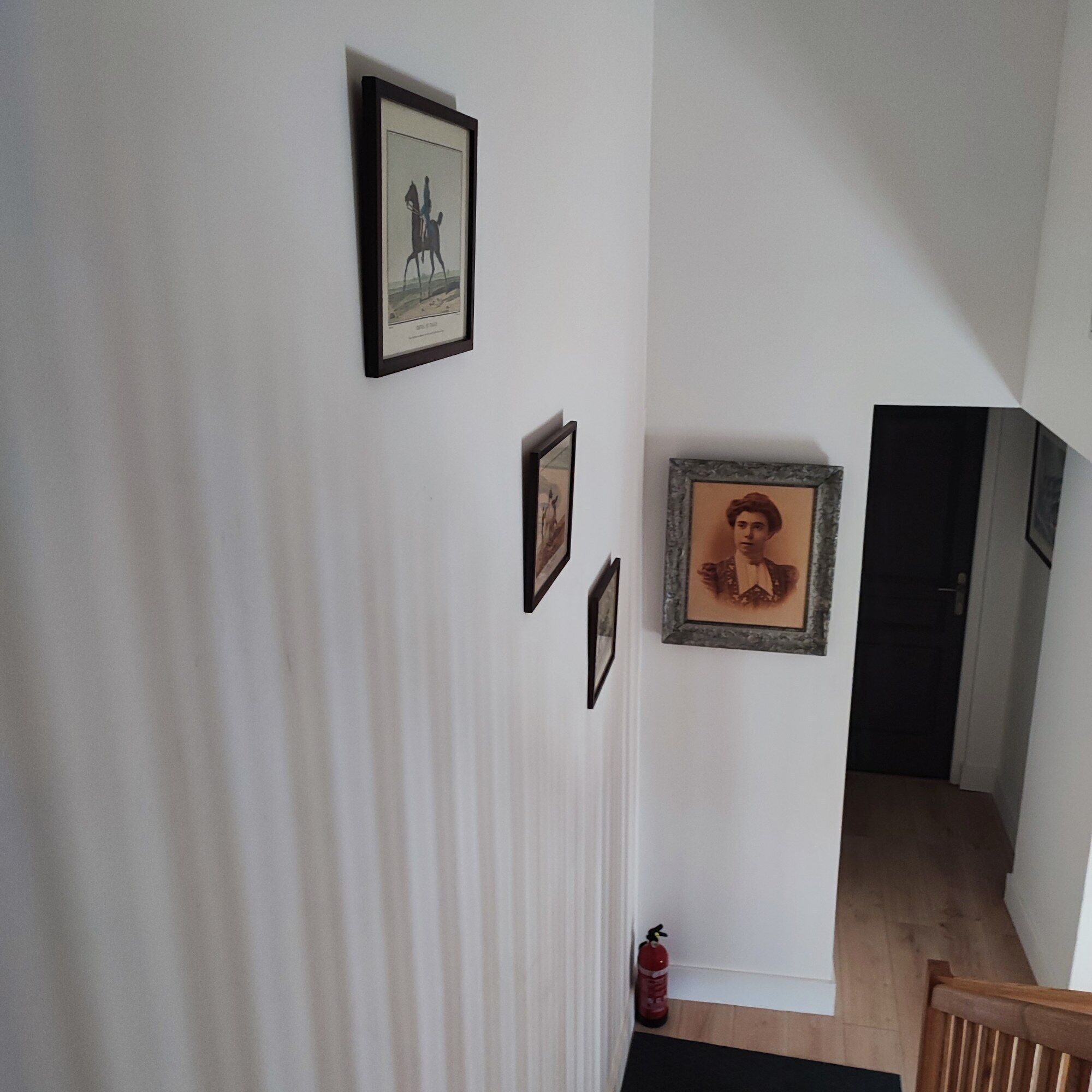A hallway with framed pictures on the wall, a fire extinguisher, and a wooden staircase.